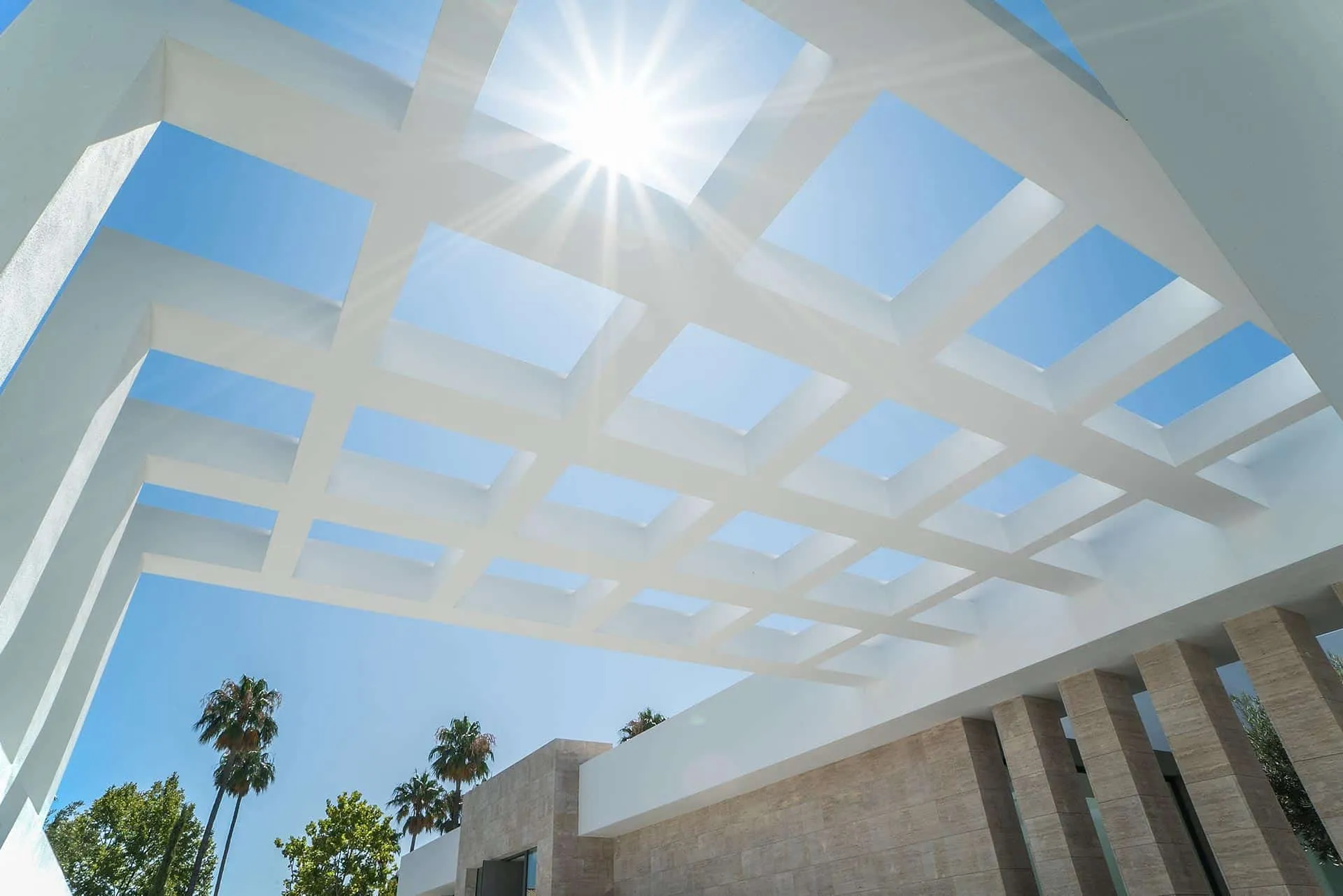Modern white geometric pergola design with sun rays filtering through, highlighting natural stone walls and clear blue sky.