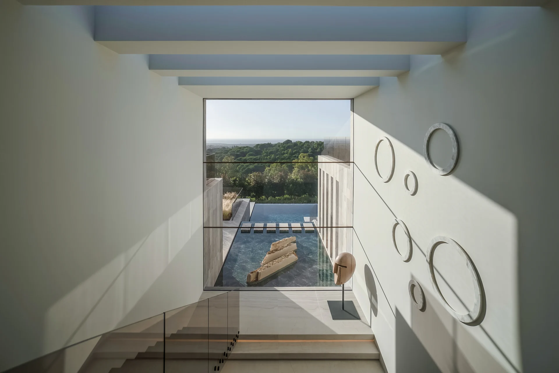 The internal staircase at Villa ENSO features a large window framing sculptures set against pools of water, highlighting the interplay of natural light and the design by ARK Architects.